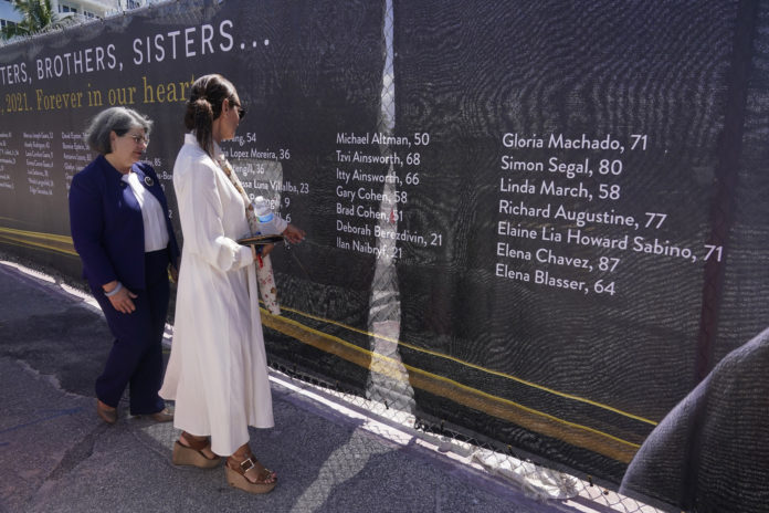 Ronit Naibryf, right, shows Miami-Dade County Mayor Daniella Levina Cava, the name of her son Ilan Naibryf, Thursday, May 12, 2022, in Surfside, Fla. A large banner with the names of the 98 people killed in the collapse of the Champlain Towers South condominium nearly a year ago, was installed around the site. (AP Photo/Marta Lavandier)